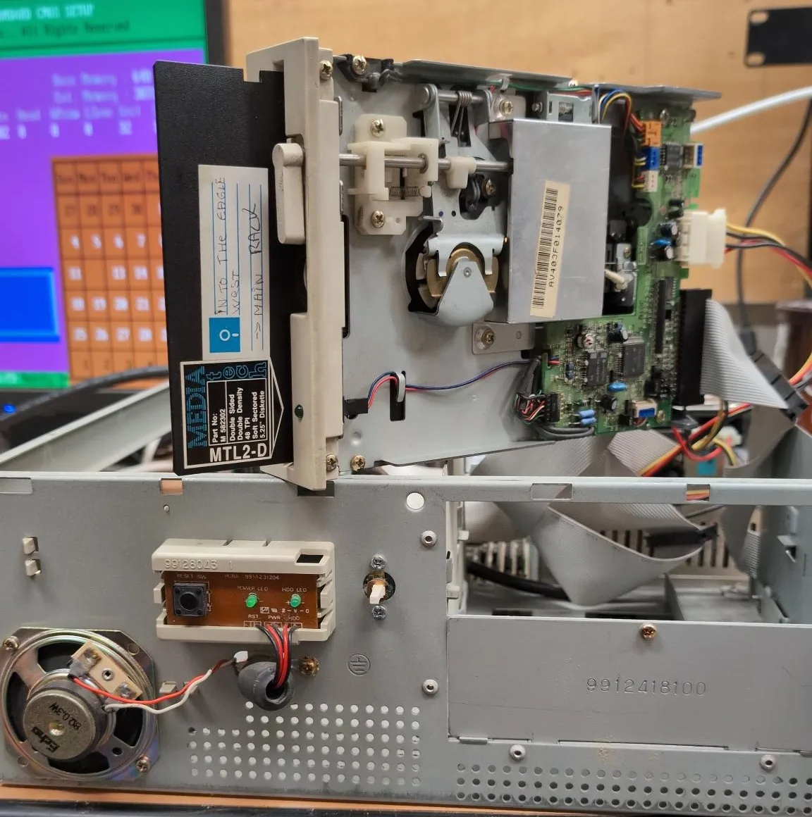 Lecteur de disquette 5 pouces 1/4 ouvert, mécanisme et carte électronique apparents, posé sur le châssis d'un ancien PC avec une disquette insérée